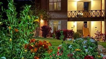Un edificio de dos plantas con un balcón, rodeado por un jardín con diversas flores.