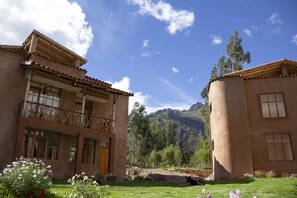 Un edificio de dos pisos con balcón, rodeado de un jardín cuidado y montañas al fondo.