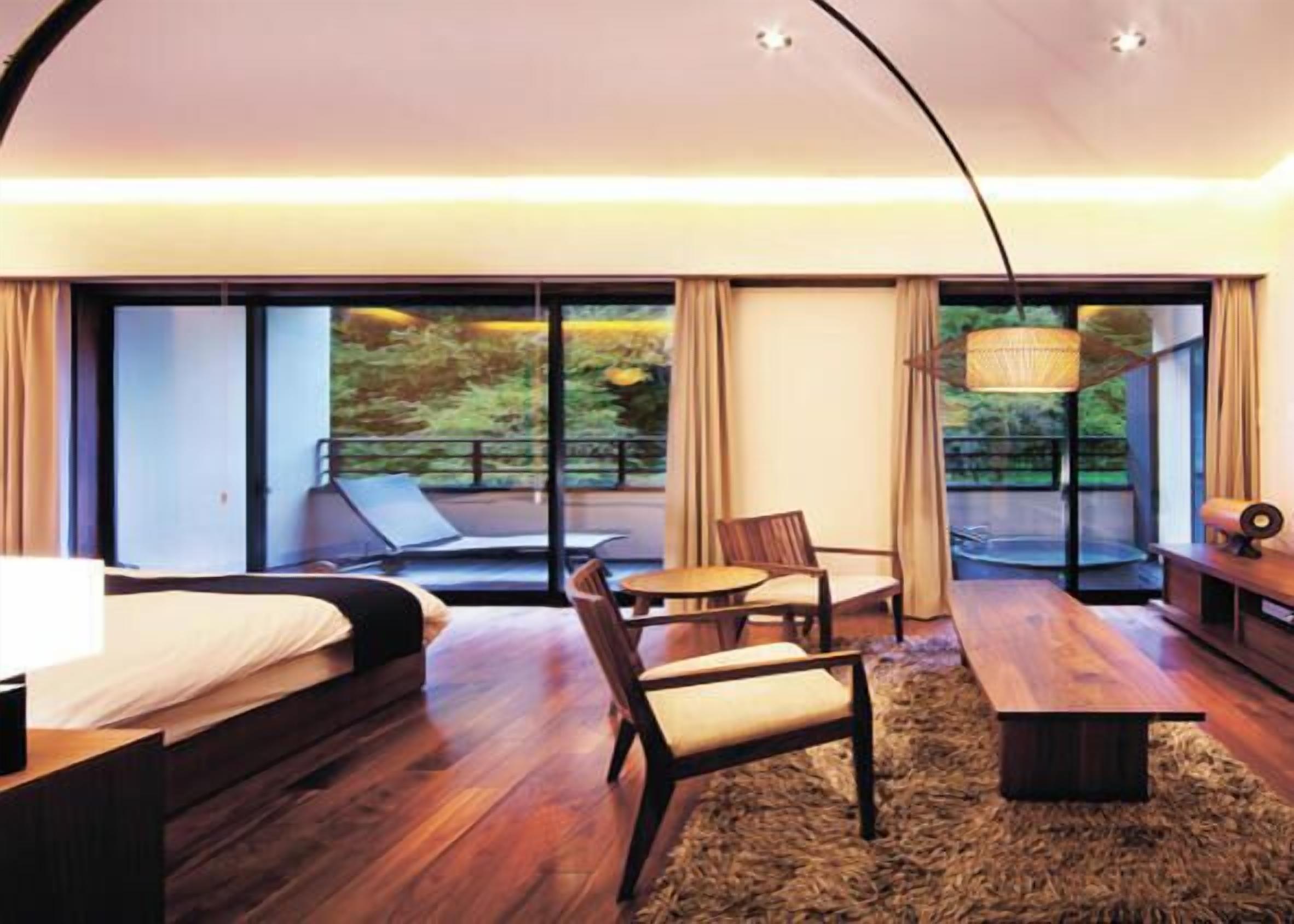 A modern hotel room with a bed, a desk, a chair, a small table, and a large window offering a view of greenery.