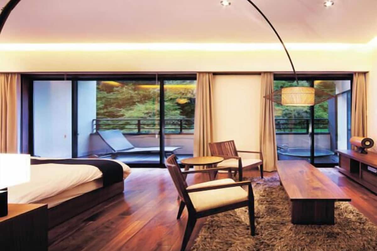 A modern hotel room with a bed, a desk, a chair, a small table, and a large window offering a view of greenery.