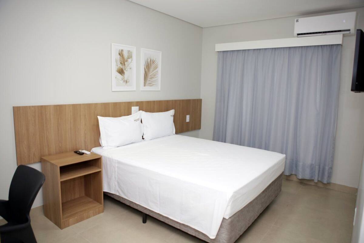 Une chambre d’hôtel avec une tête de lit en bois, du linge de lit blanc, une petite table de chevet en bois, une télécommande et un climatiseur fixé au mur.