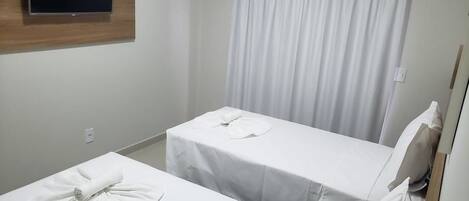 Quarto de hotel com duas camas de solteiro, uma televisão de tela plana presa a um painel de madeira, cortina branca e aparelho de ar condicionado branco.