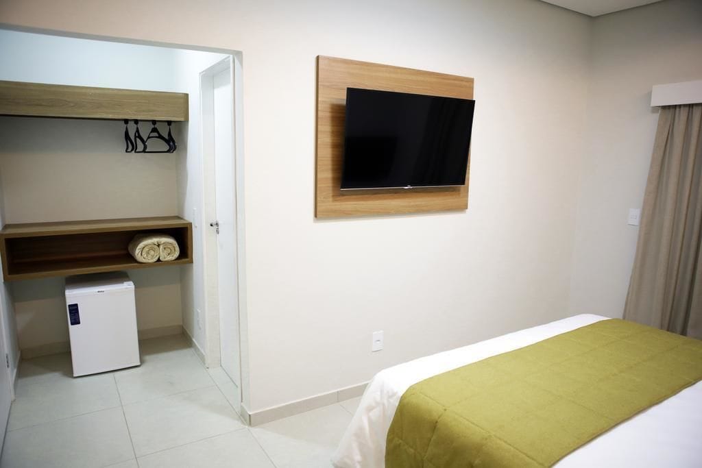 Habitación de hotel con cama, televisor montado en la pared, un refrigerador pequeño y un estante de madera con toallas.