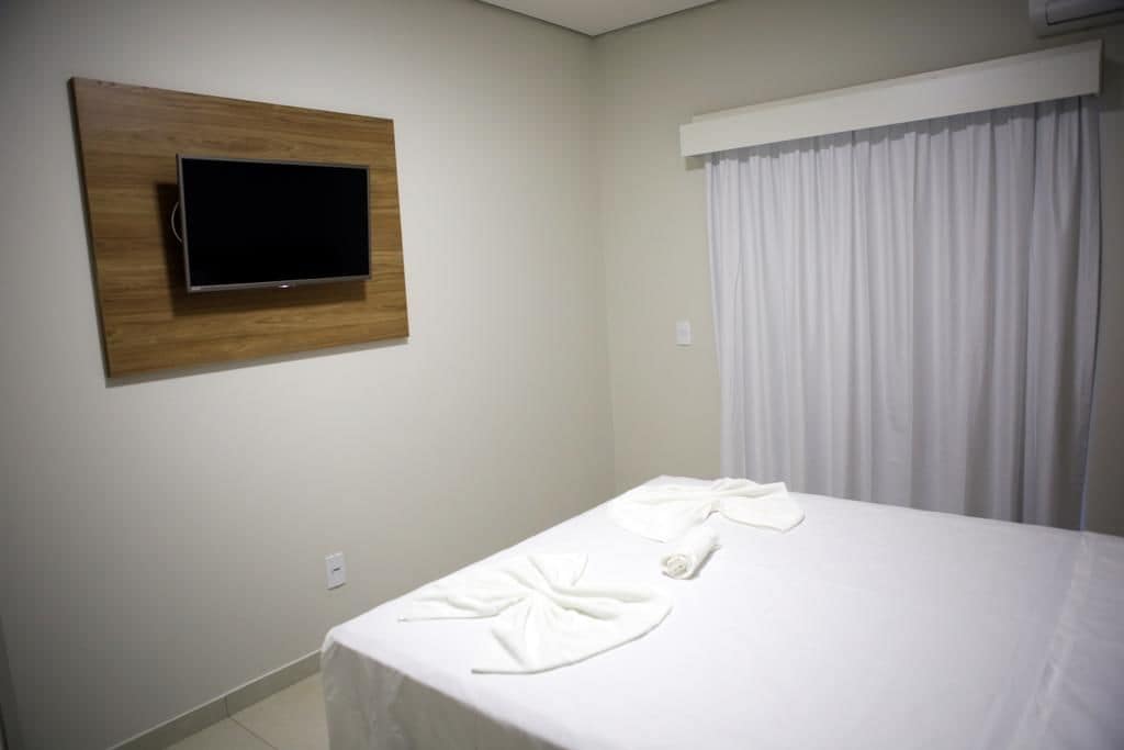 A hotel room with a bed, a flat-screen TV mounted on a wooden panel, and a window with white curtains.