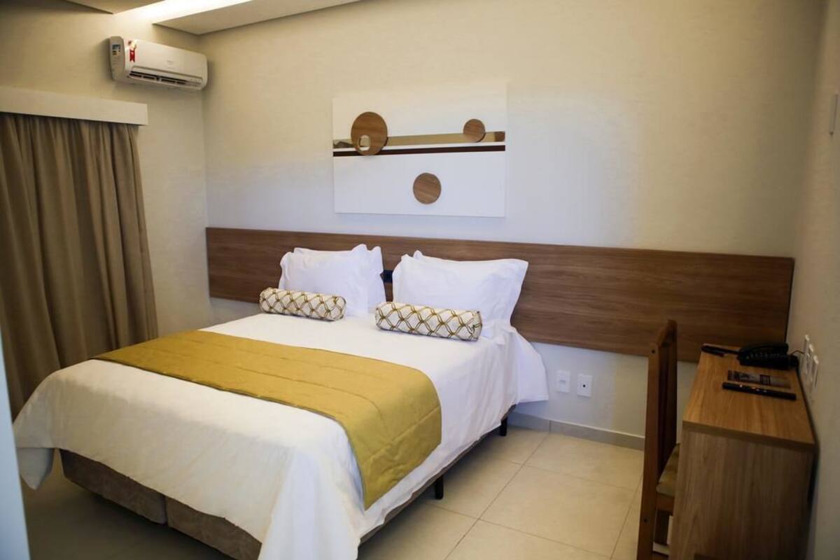 Une chambre d’hôtel avec un lit, une tête de lit en bois, une table de chevet et un climatiseur fixé au mur.