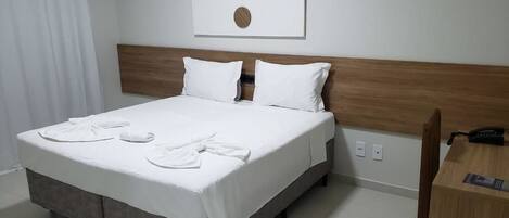 Una cama bien tendida con sábanas blancas y dos almohadas, cabecero de madera, una obra de arte colgada en la pared y un escritorio de madera con un teléfono.
