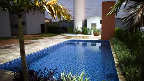Ein Schwimmbecken mit gefliesten Rändern, umgeben von Grünflächen und einem modernen Gebäude mit zylindrischer Struktur.