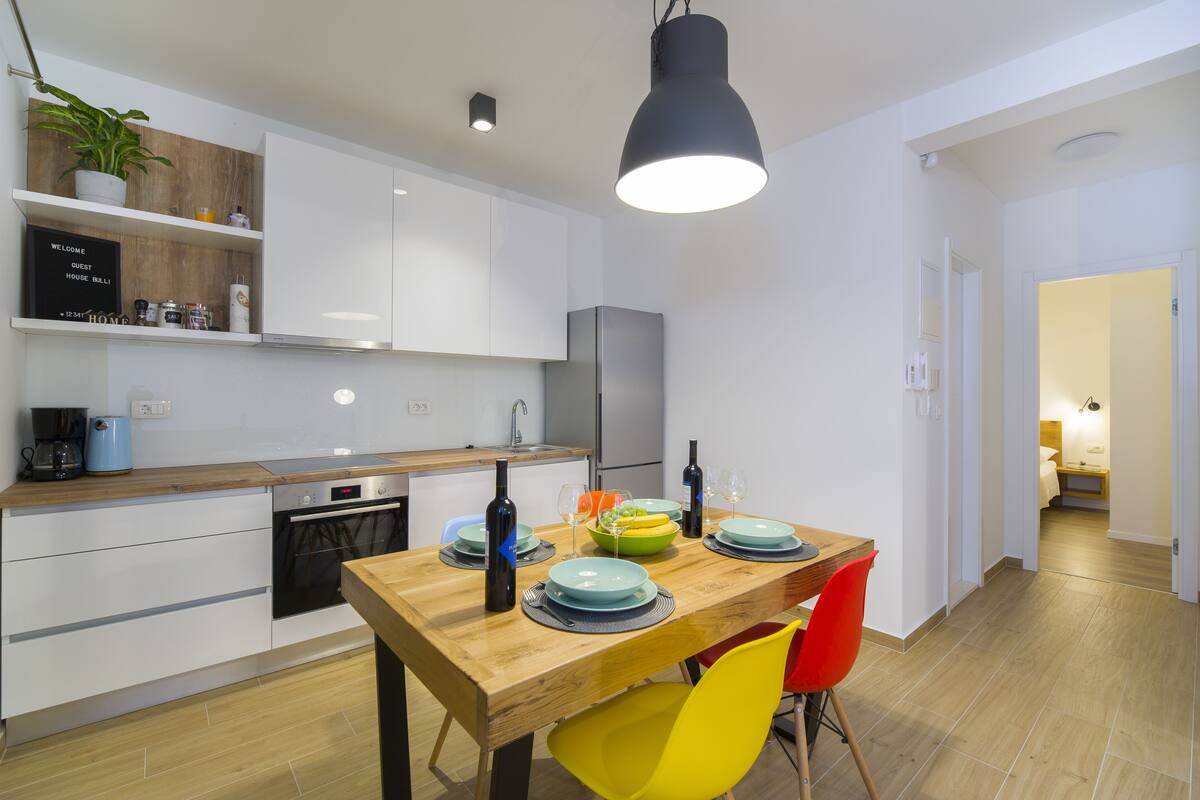 A modern kitchen with white cabinets, a wooden countertop, and a dining table set with plates, glasses, and a bottle.