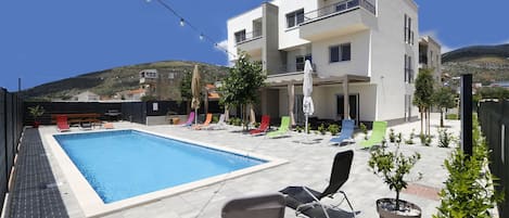 A modern building with a swimming pool, outdoor seating, and a hillside backdrop.