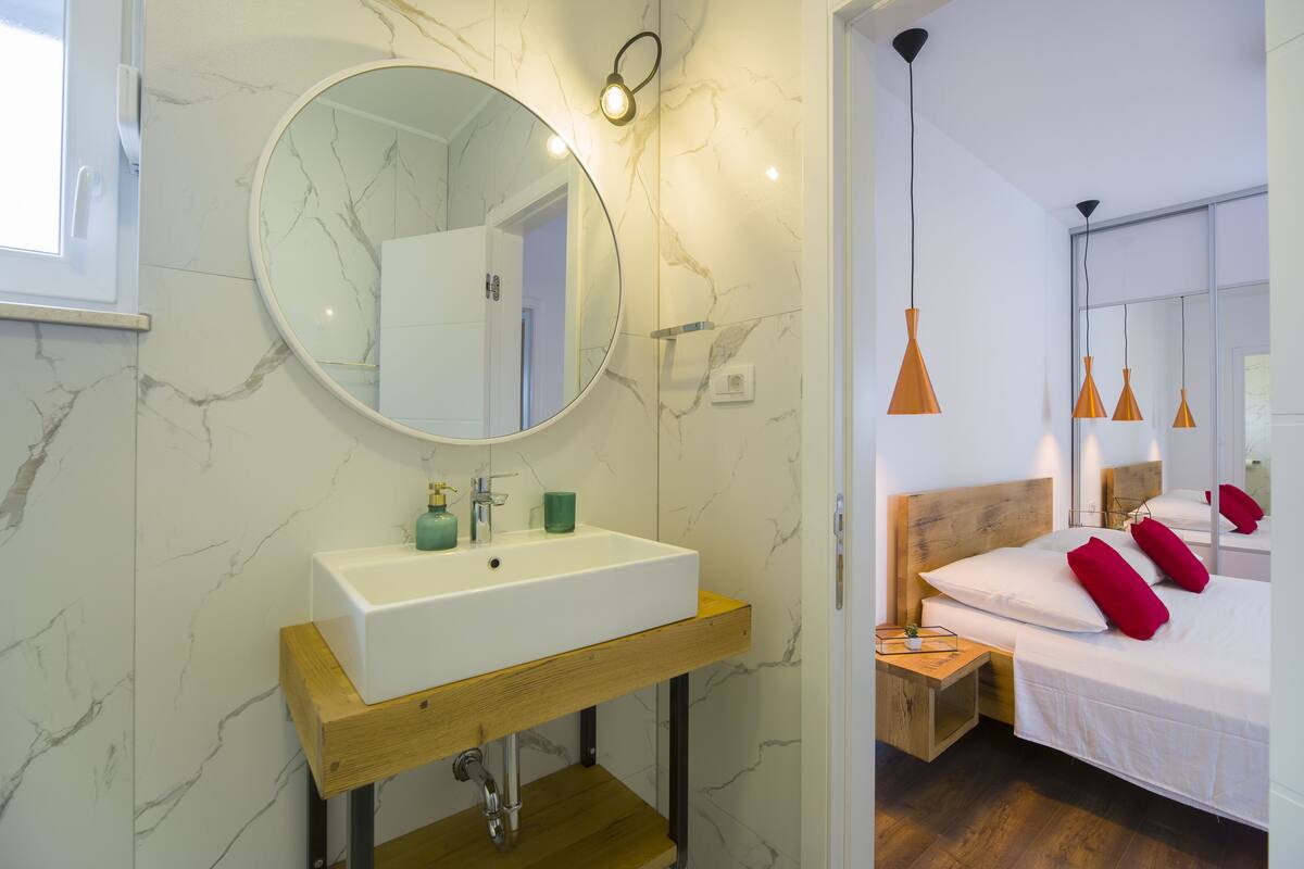 A modern bathroom with a marble wall, a wooden vanity, and a large round mirror.