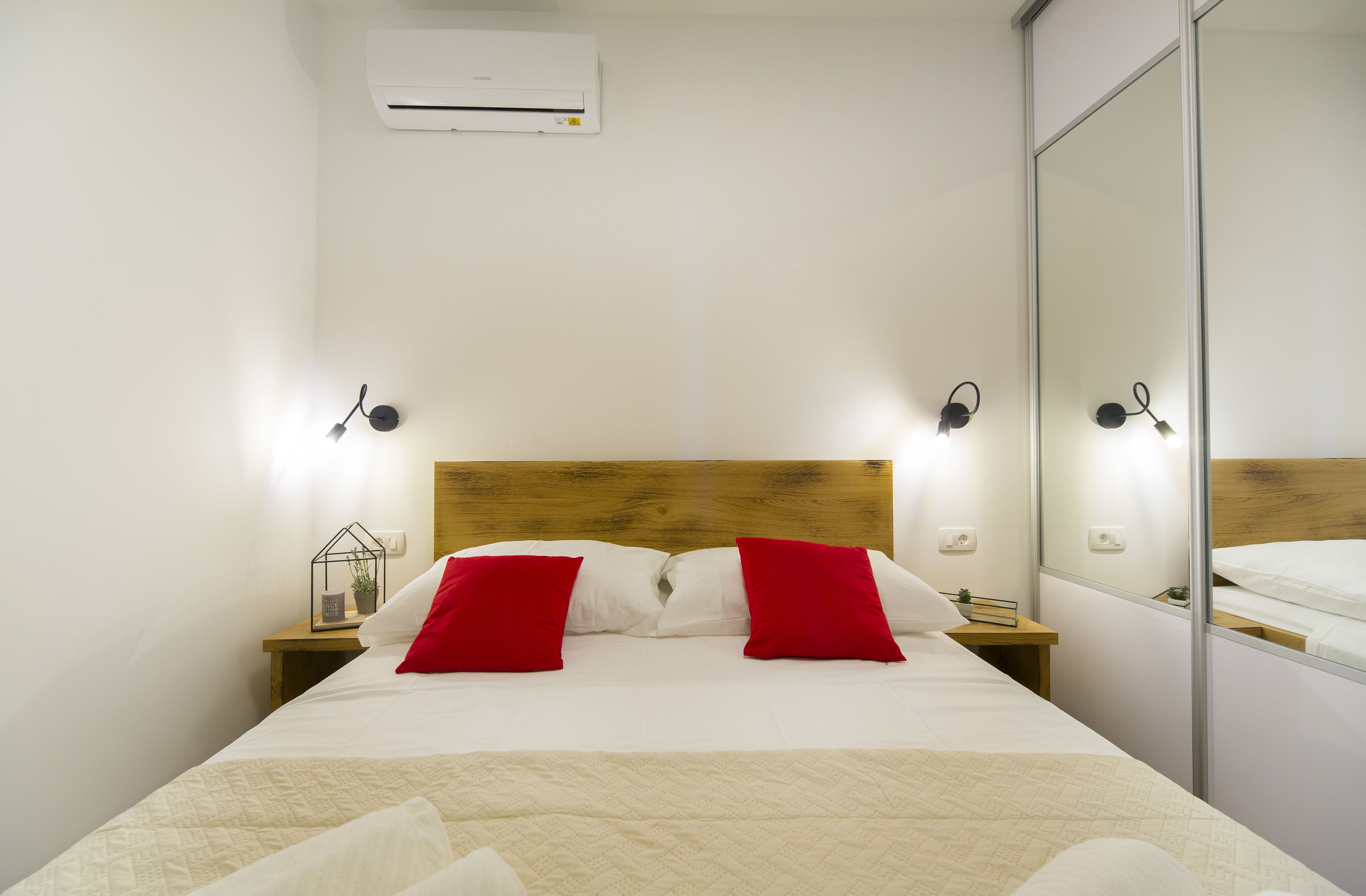 Una cama bien hecha con almohadas rojas, un cabecero de madera, mesitas de noche con lámparas y un espejo grande en la pared.
