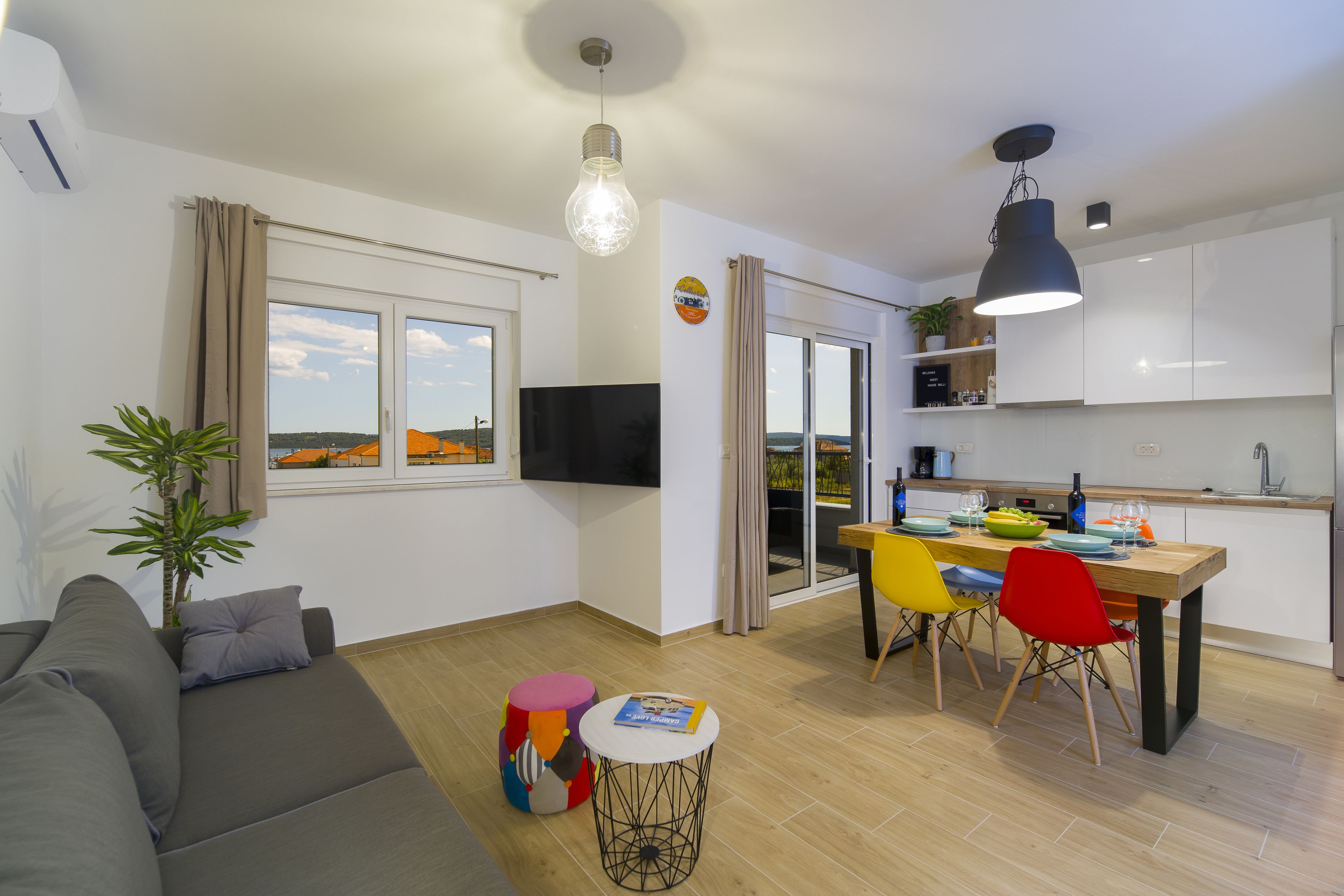 Ein modernes Wohnzimmer mit einer Couch, einem kleinen runden Tisch und einem farbenfrohen Hocker. Es gibt einen Essbereich mit einem Holztisch und Stühlen.