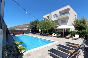 Une piscine entourée de chaises longues et de parasols, le tout bordé par un bâtiment doté d’un balcon.