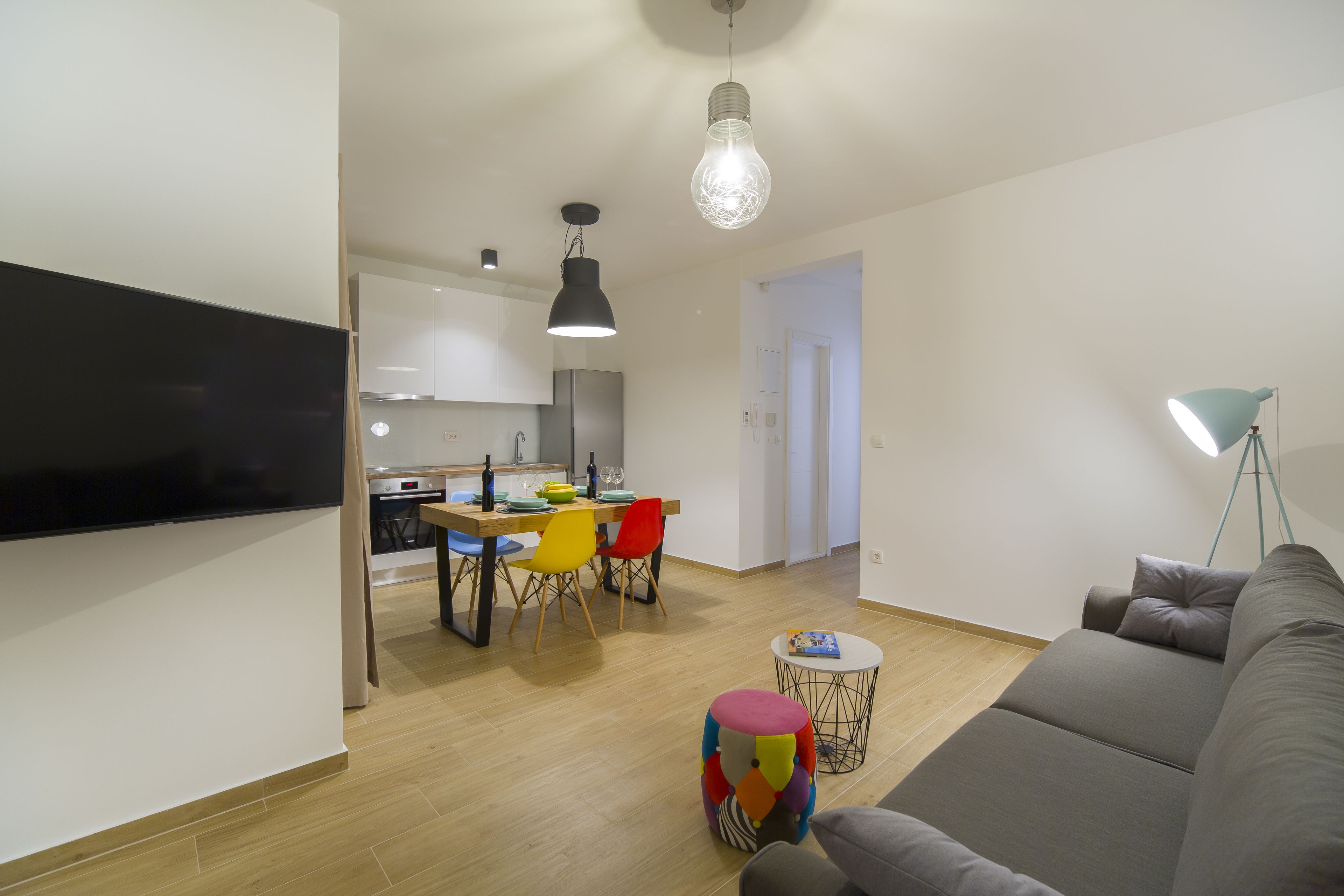 A modern living room with a gray sofa, a colorful ottoman, and a dining area with a wooden table and colorful chairs.