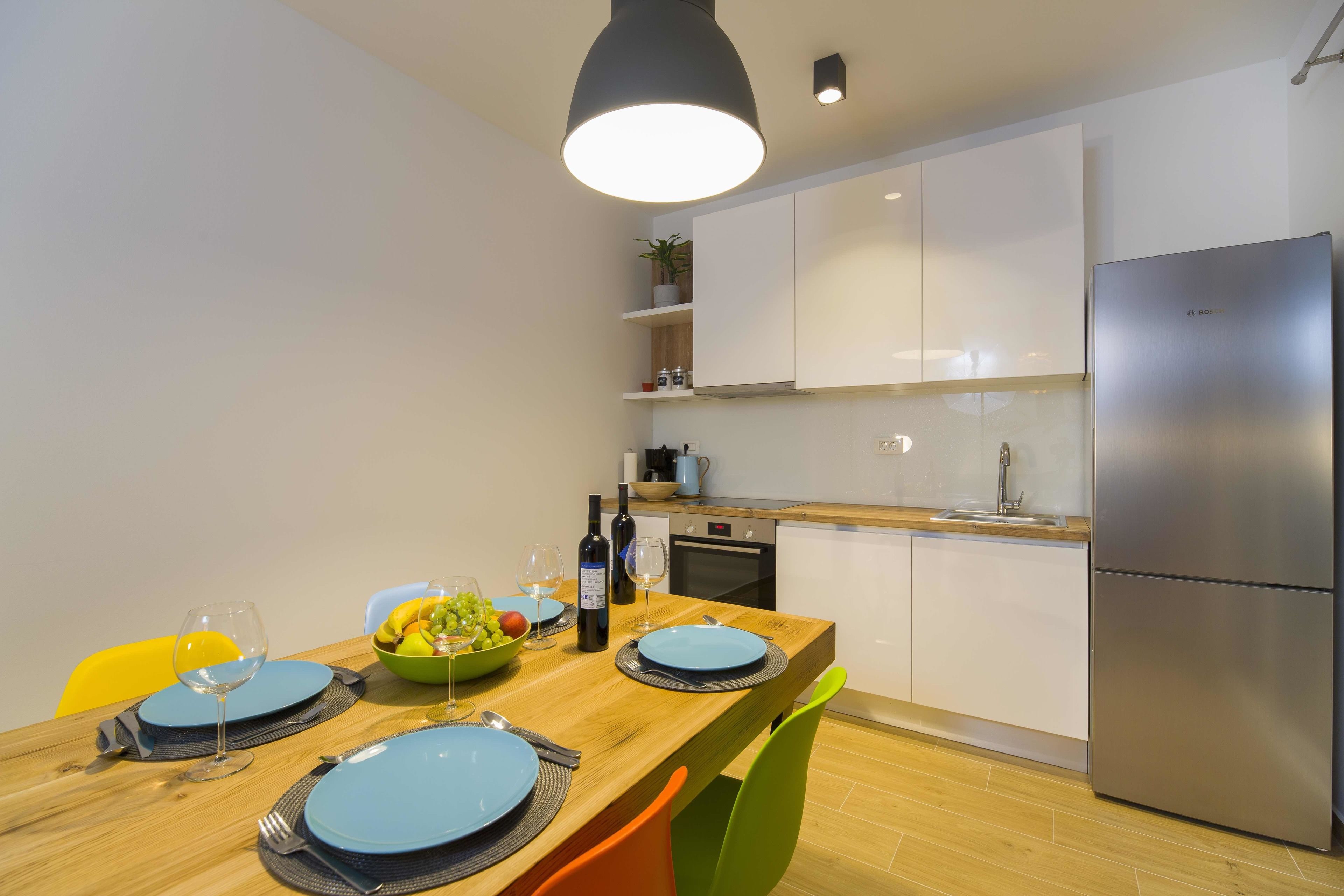 A modern kitchen with white cabinets, a stainless steel refrigerator, and a wooden dining table set with blue plates and wine glasses.