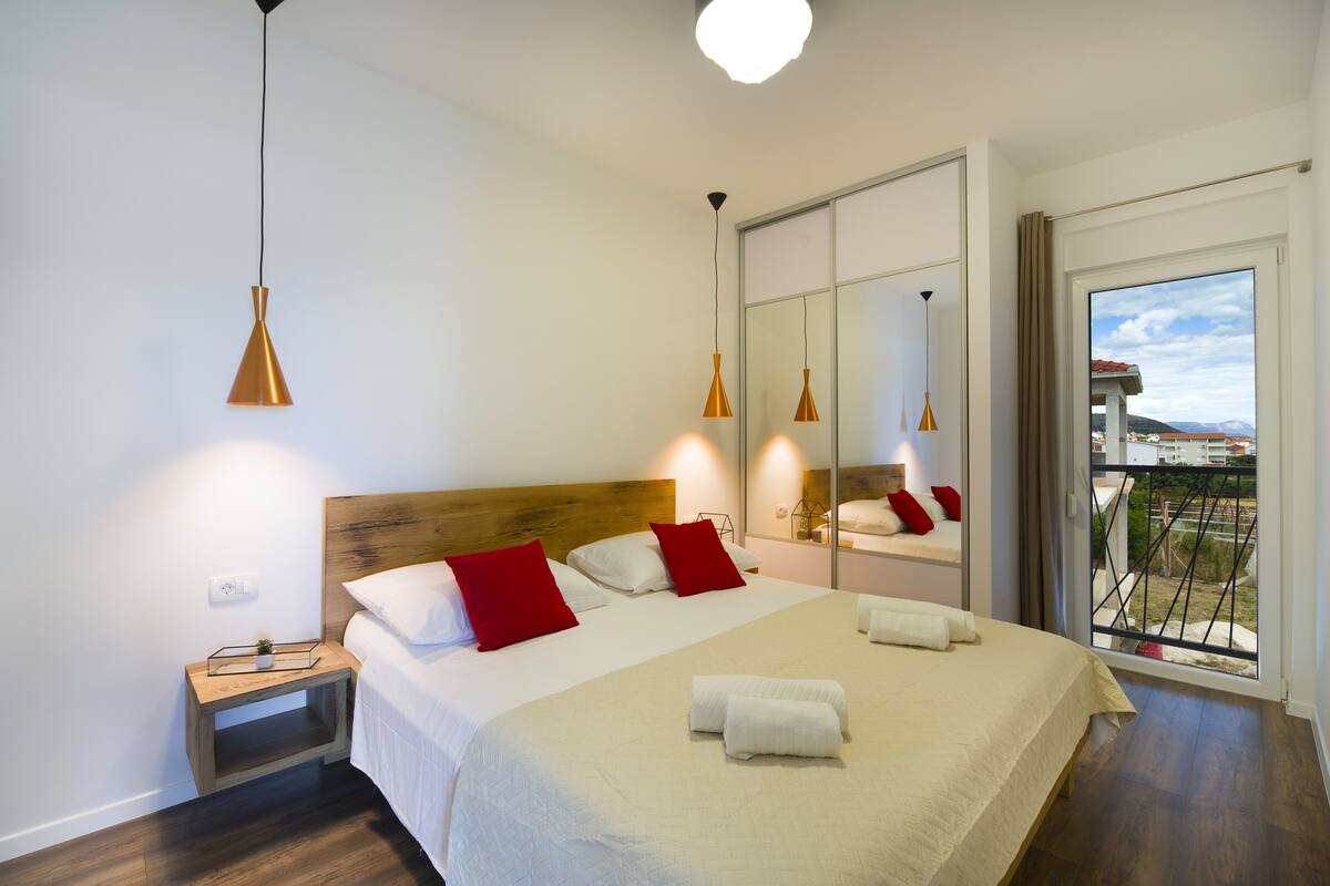 A modern hotel room with a large bed, wooden headboard, red pillows, and a balcony with a view.