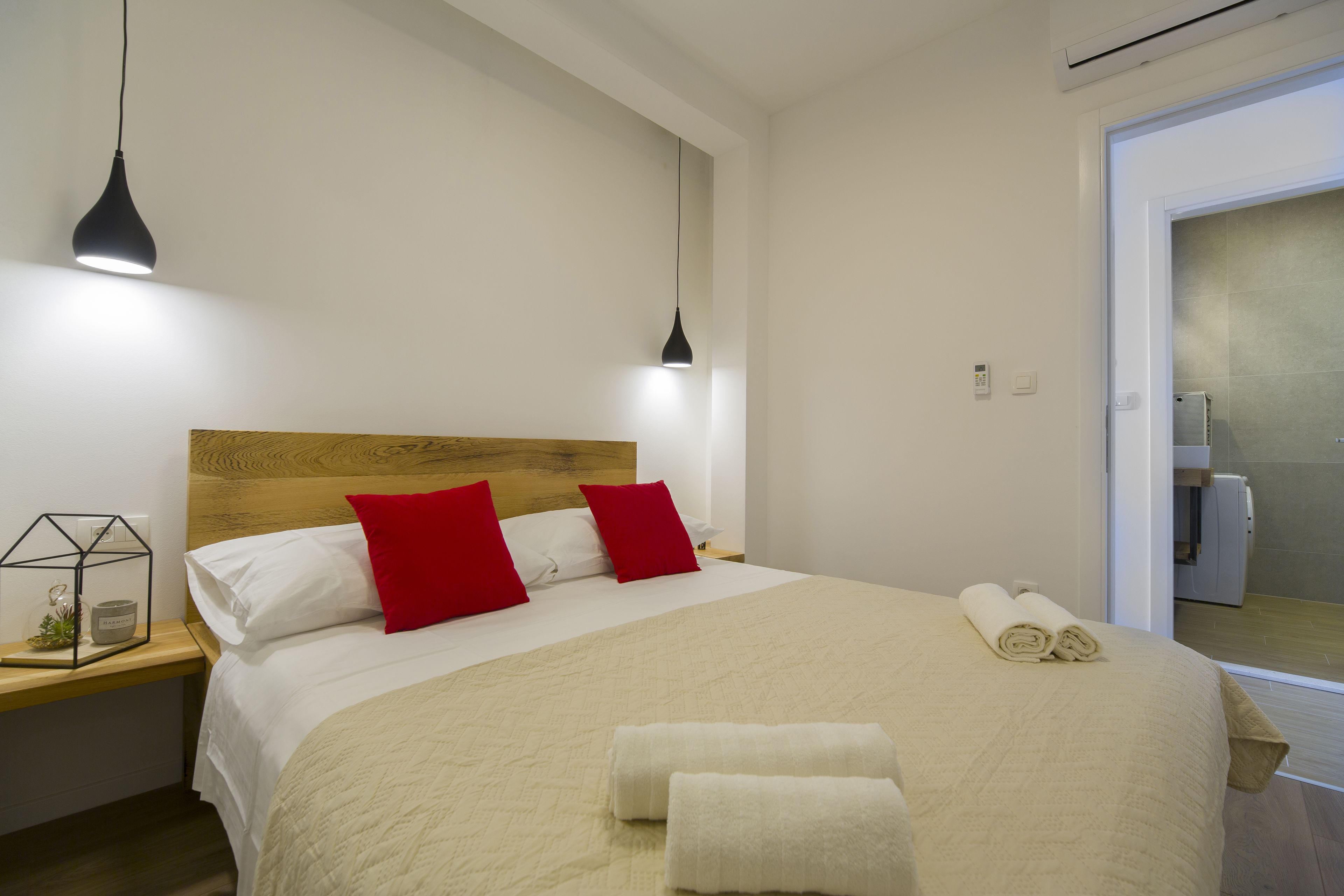 A neatly made bed with red and white pillows, a wooden headboard, and two bedside lamps. There are rolled towels on the bed and a small plant on the nightstand.