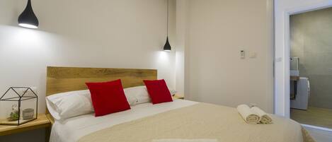 A neatly made bed with red and white pillows, a wooden headboard, and two bedside lamps. There are rolled towels on the bed and a small plant on the nightstand.
