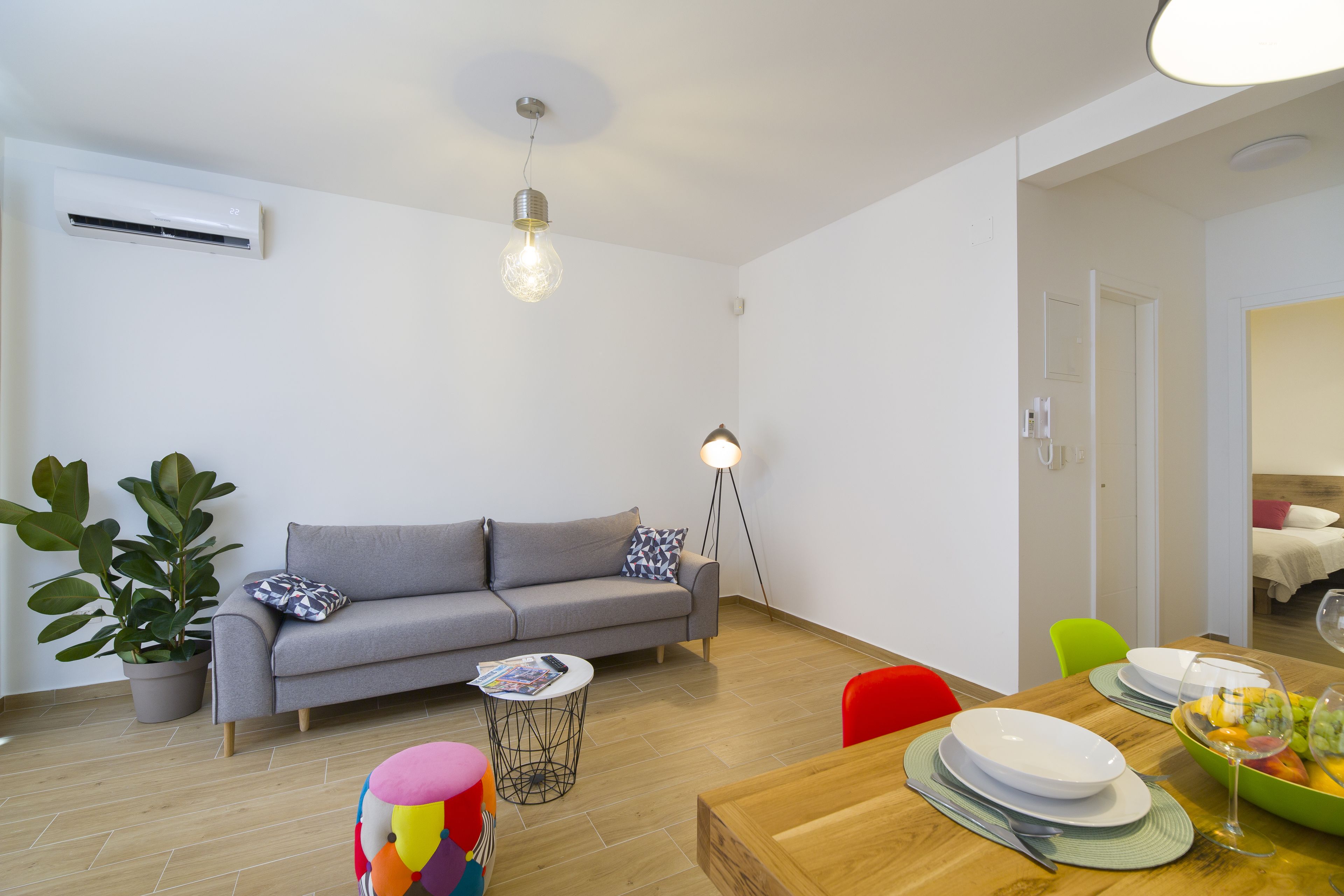 A modern living room with a grey sofa, a wooden dining table set with plates and glasses, a floor lamp, and a potted plant.