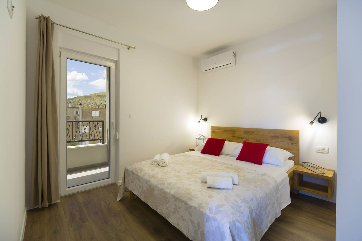 A bedroom with a bed, a nightstand, a wall-mounted air conditioner, and a view through a sliding door.