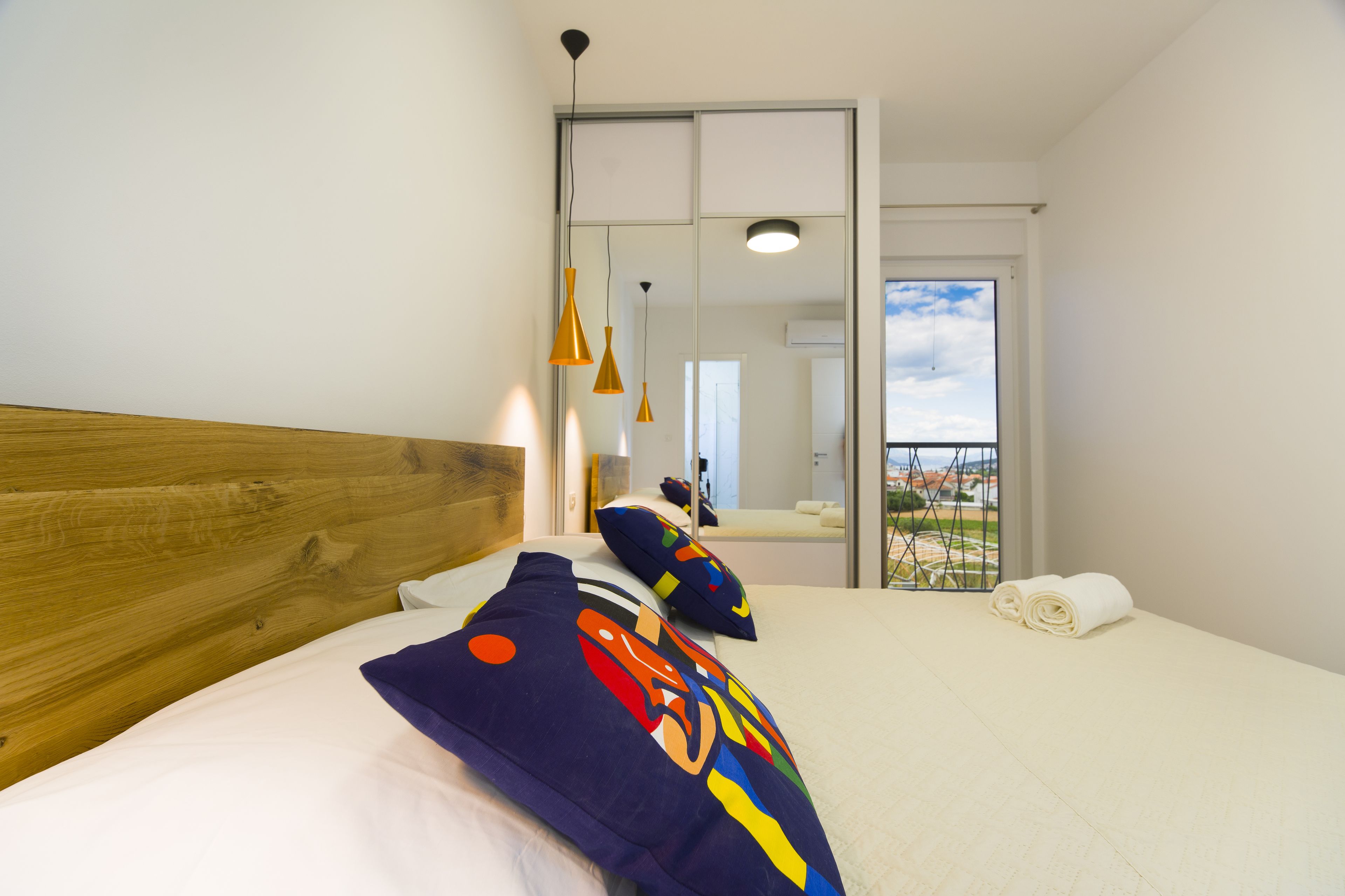 A hotel room with a wooden headboard, a large bed with white bedding, colorful pillows, a small table with a lamp, and a view of a cityscape through a balcony.