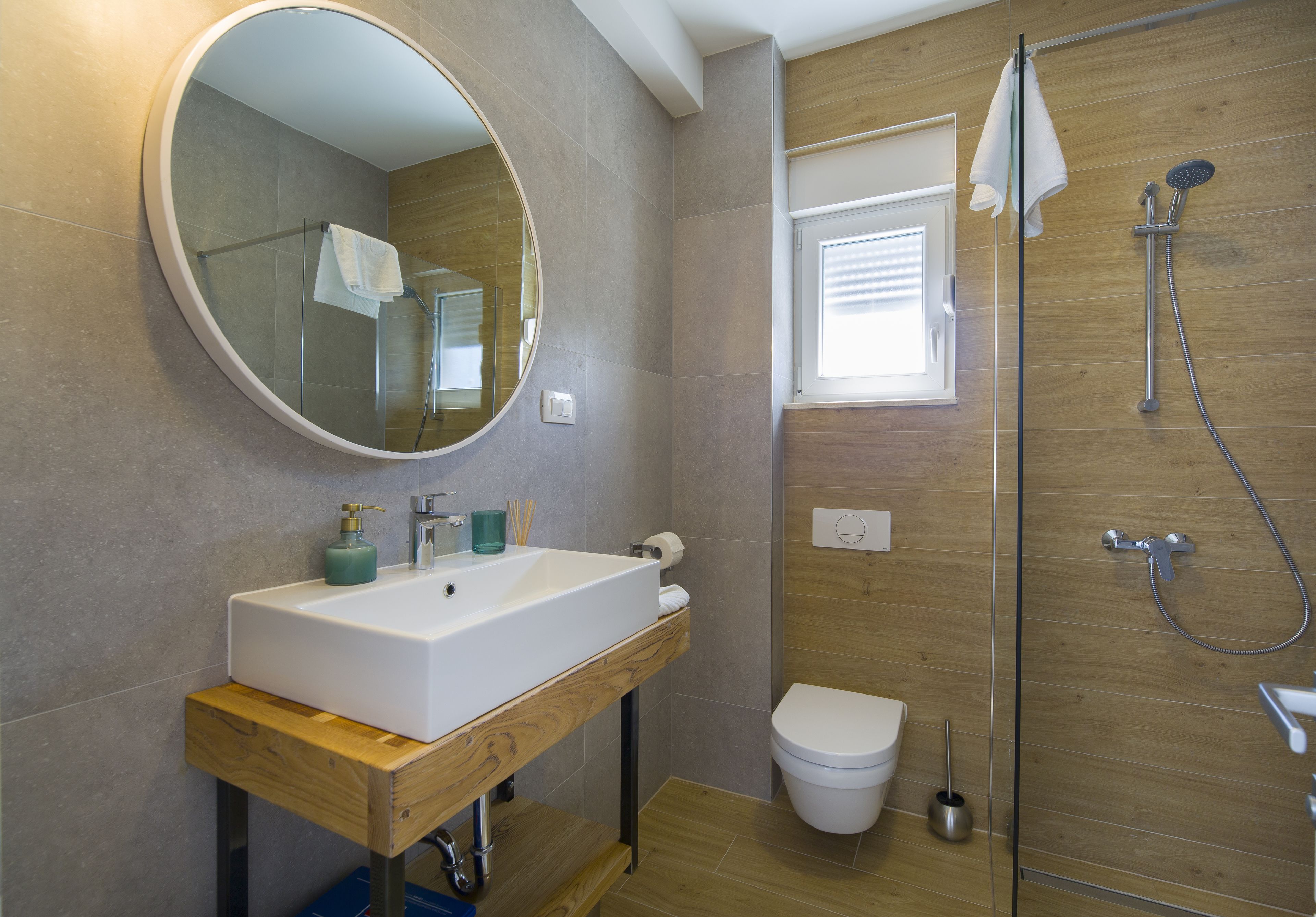 A modern bathroom with a large round mirror, a white sink, a toilet, and a shower area.