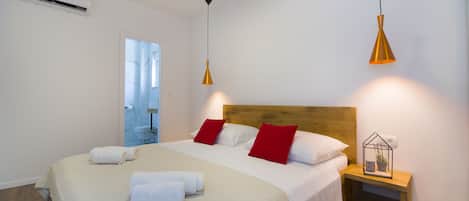 Een opgemaakt bed met witte en rode kussens, een houten hoofdbord en een nachtkastje met een kleine plant.
