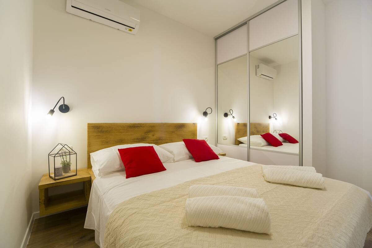 A neatly made bed with white and red pillows, a bedside table with a lamp, and a mirror reflecting the room.