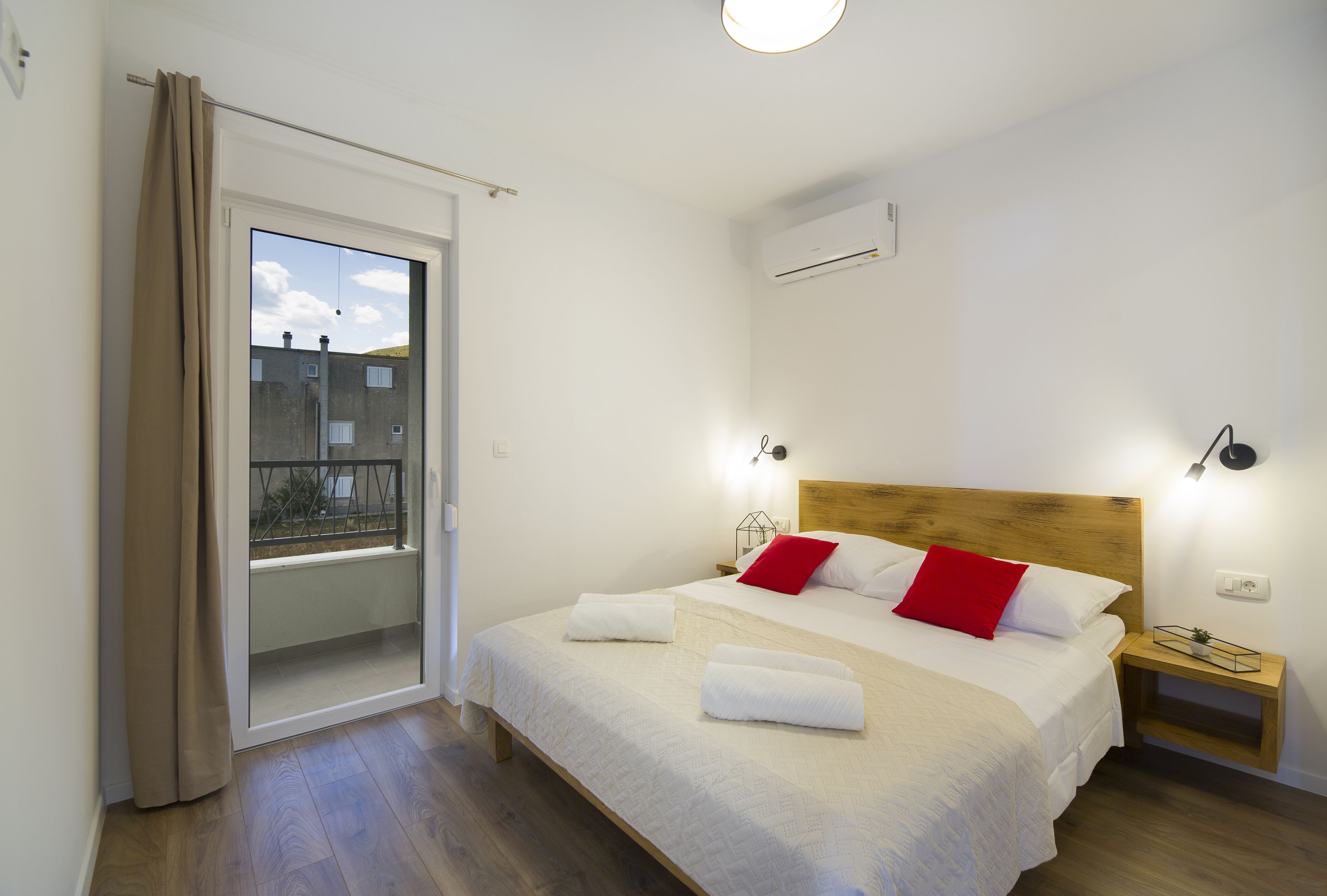 Quarto com uma cama grande, mesa de cabeceira, janela com vista para prédios e ar condicionado.