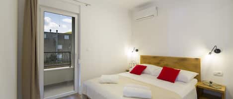 Quarto com uma cama grande, mesa de cabeceira, janela com vista para prédios e ar condicionado.