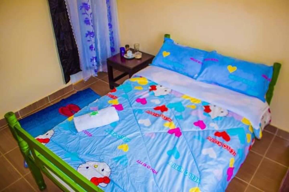 A bed with a Hello Kitty themed bedspread, a small bedside table with a lamp, and a window with curtains.