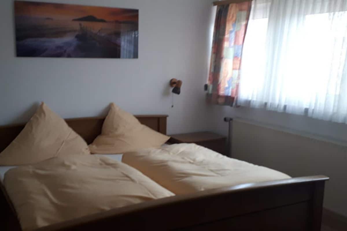 Un dormitorio con una cama, dos mesitas de noche, una lámpara de pared y un ventanal con cortinas.