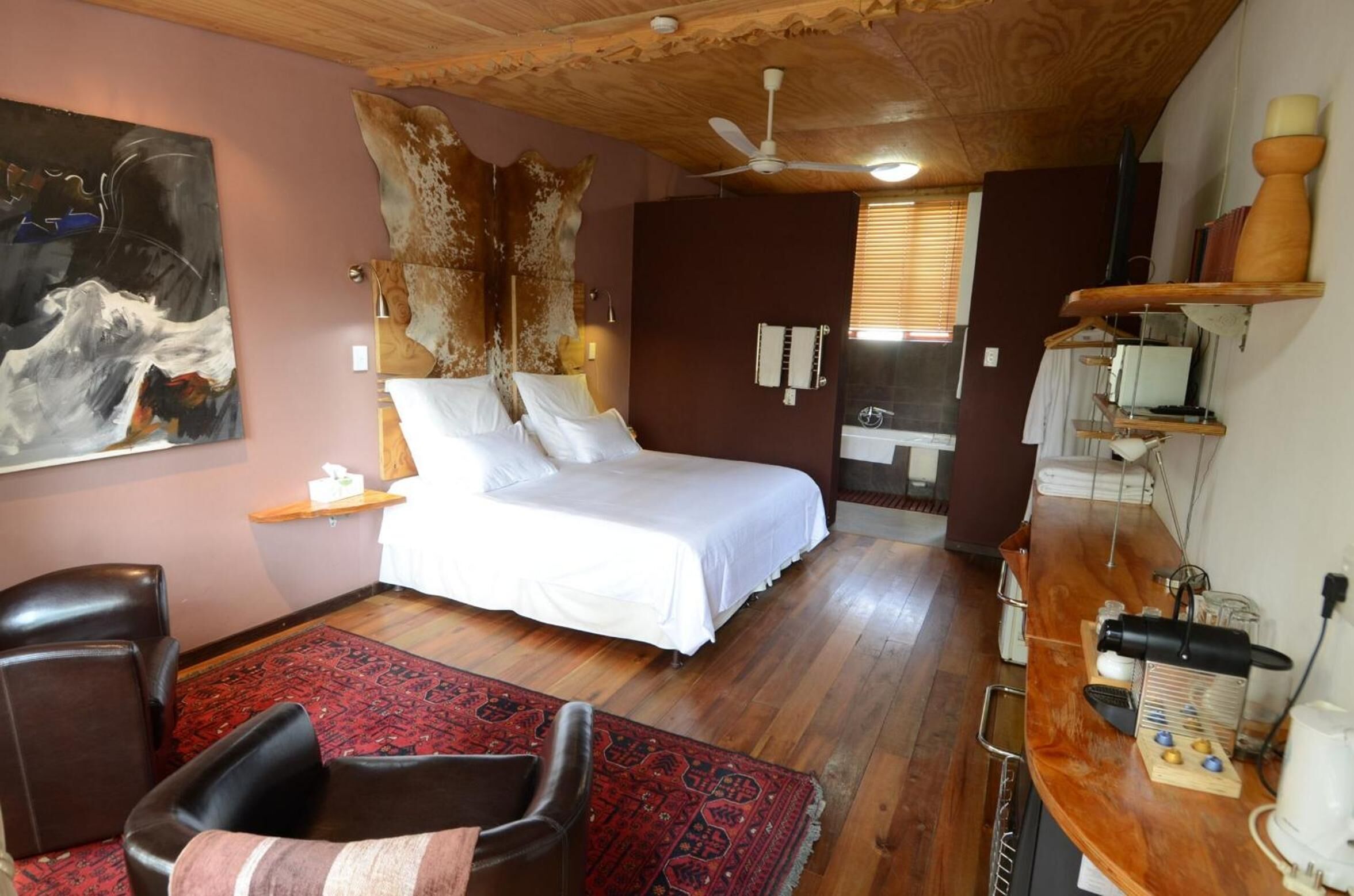 A bedroom with a bed, a wooden ceiling, a ceiling fan, a red rug, a wooden desk with a lamp, and a painting on the wall.