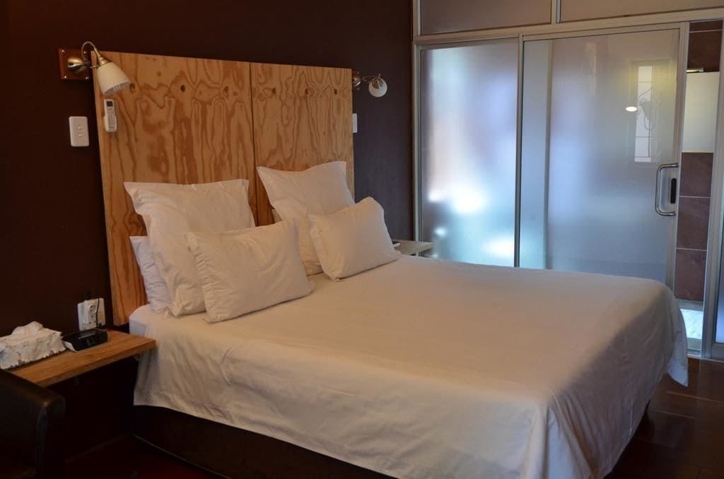 A modern hotel room with a wooden headboard, a large bed with white linens, a bedside table with a lamp, and a sliding glass door leading to a bathroom.