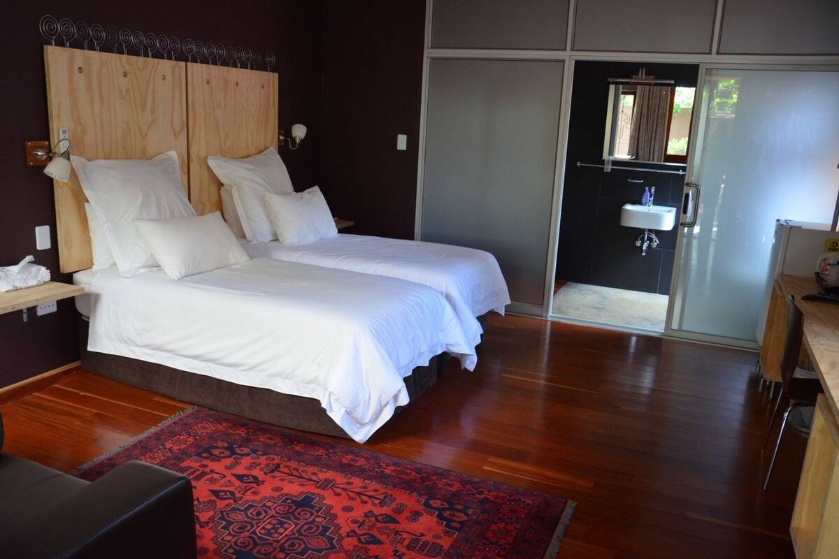 A double bed with white bedding, a wooden headboard, a red patterned rug, and a view of a bathroom with a sink and mirror.