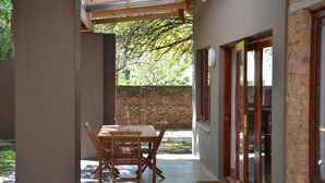 Un porche cubierto con una mesa y sillas de madera, un muro de ladrillo y una puerta de cristal que da a un jardín.