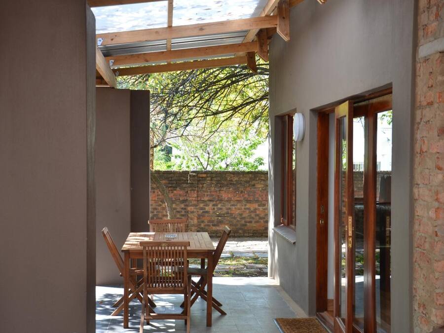 A covered patio with a wooden table and chairs, a brick wall, and a glass door leading to a garden.