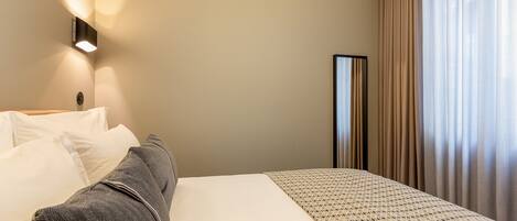 A hotel room with a bed, pillows, a bedside table, a lamp, and a window with curtains.