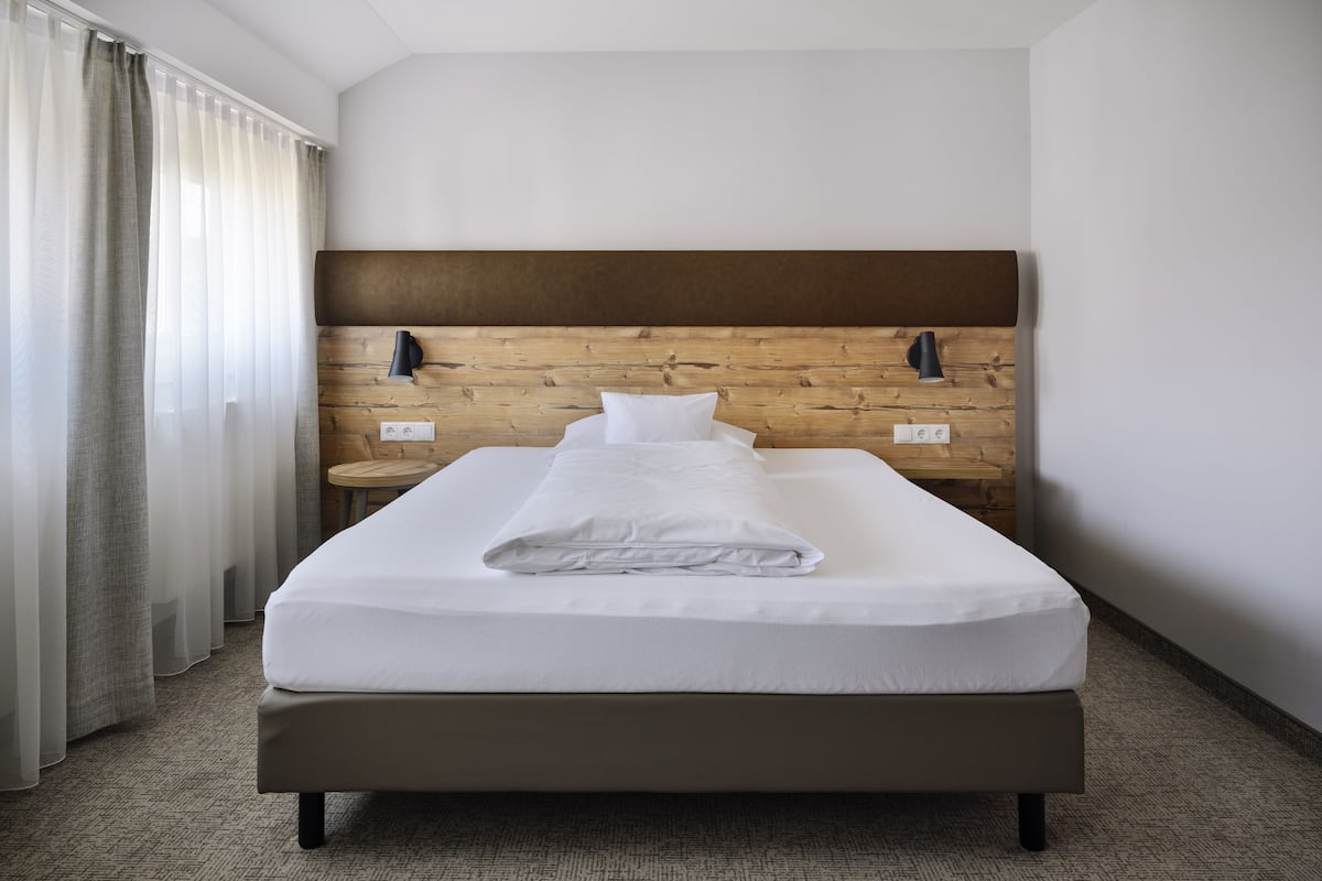 A neatly made bed in a hotel room with a wooden headboard, a small bedside table, and a wall-mounted lamp.