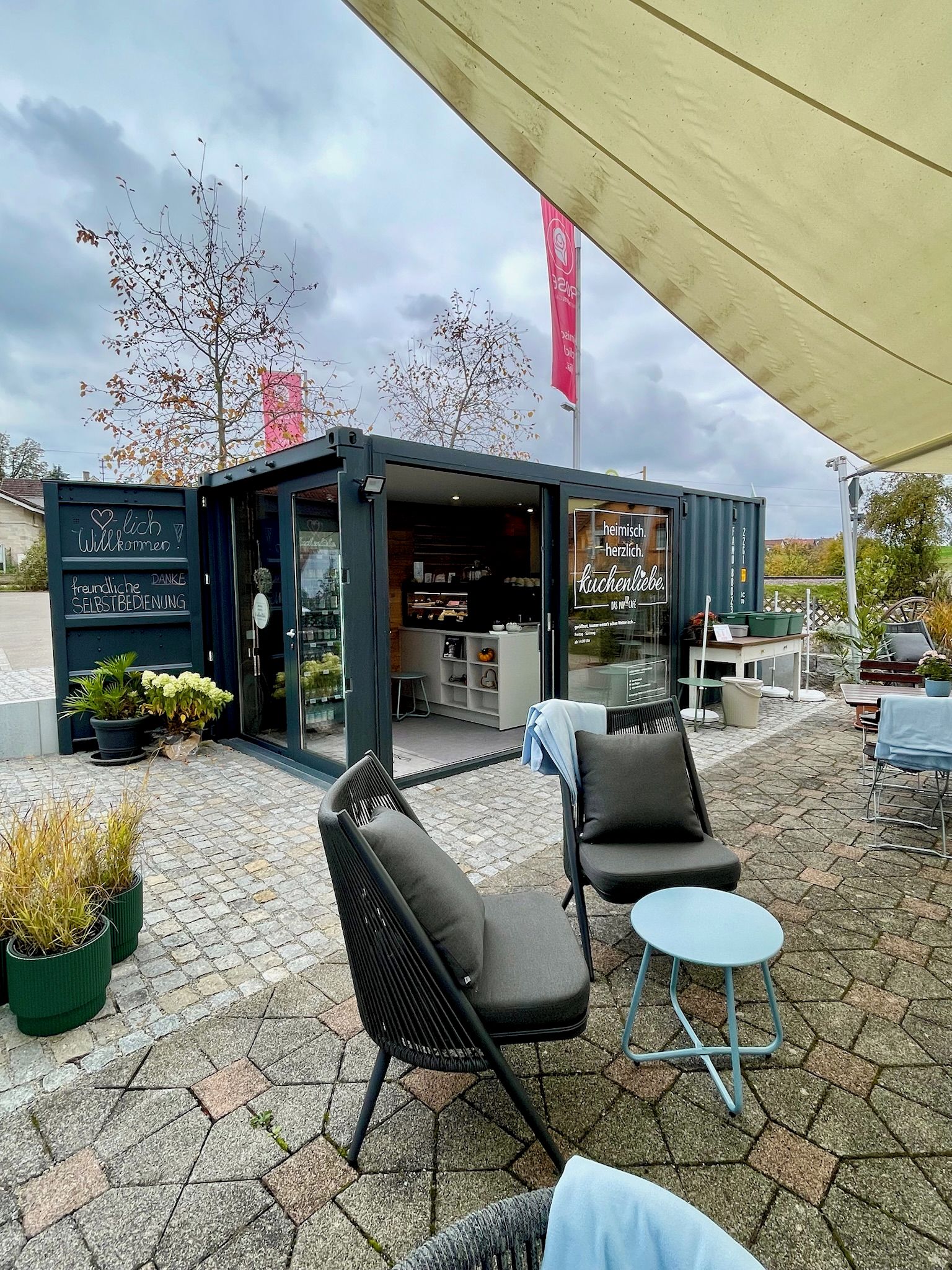 Un área de estar al aire libre con un café, que incluye sillas y mesas, bajo un toldo.
