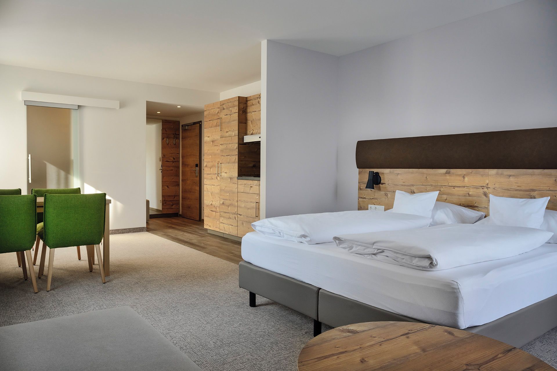 Une chambre d’hôtel moderne avec un grand lit, une tête de lit en bois, un coin salon avec des fauteuils verts et un mur d’accent en bois.