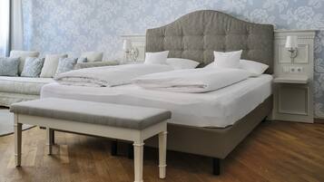 Una cama doble bien hecha con cabecera tapizada en gris, una mesita de noche con lámpara, y un banco frente a la cama.