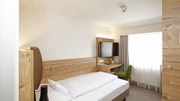 Ein Hotelzimmer mit einem Holz-Kopfteil, einem Bett mit weißer Bettwäsche, einem Schreibtisch mit Computer, einem grünen Stuhl und einem Fenster mit Vorhängen.