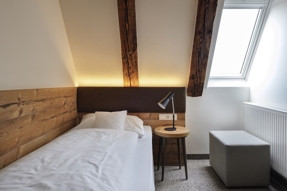 A hotel room with a wooden headboard, a small round table with a lamp, a grey ottoman, and a window with natural light.