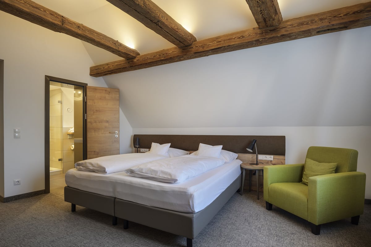 A bedroom with a large bed, a green armchair, and a wooden ceiling beam.