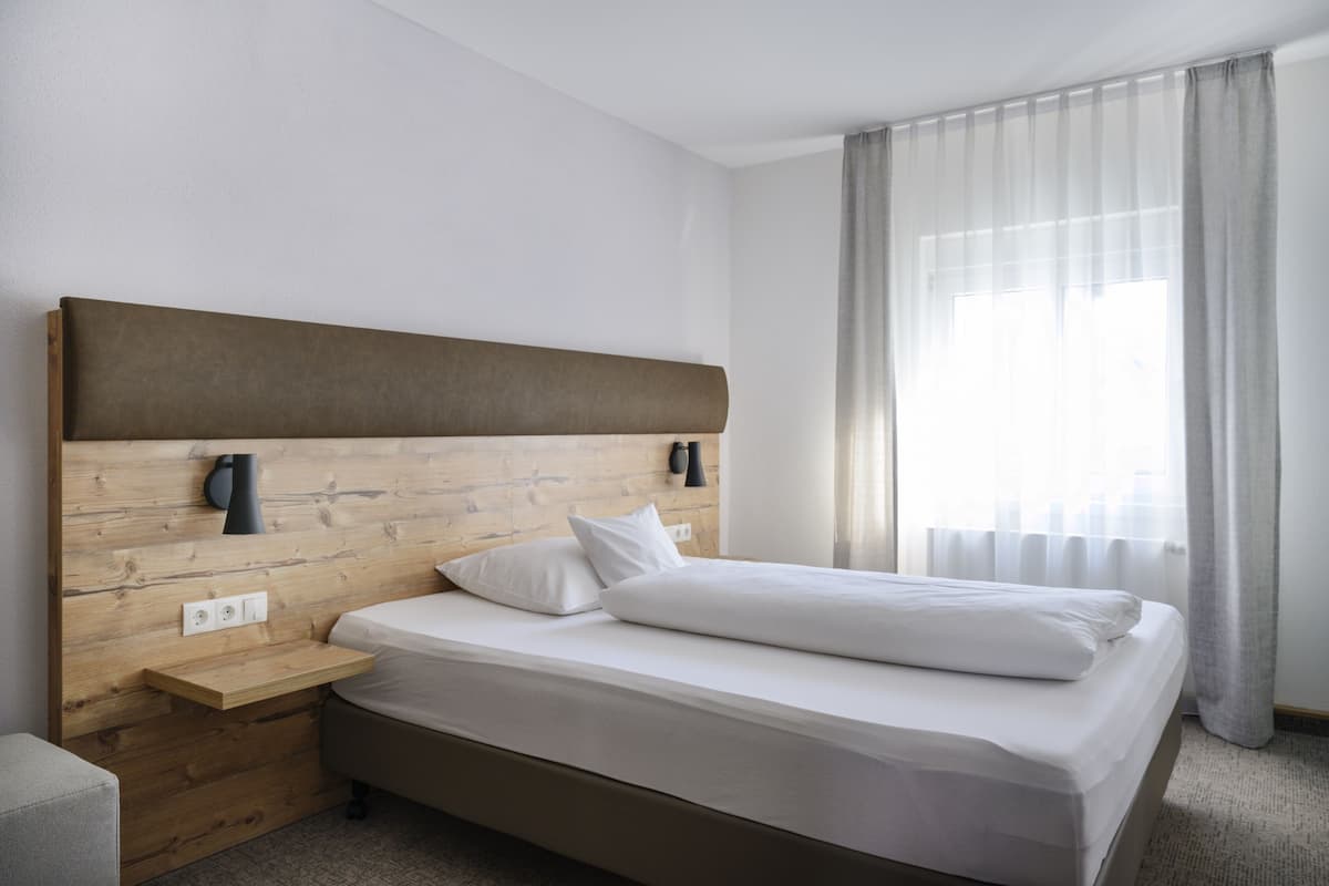 A modern hotel room with a wooden headboard, a bedside table, a bed with white linens, and a large window with sheer curtains.