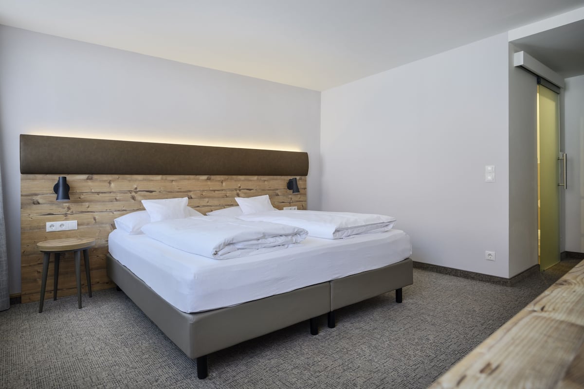 A modern hotel room with a wooden headboard, a single bed, a small table, and a bedside lamp.