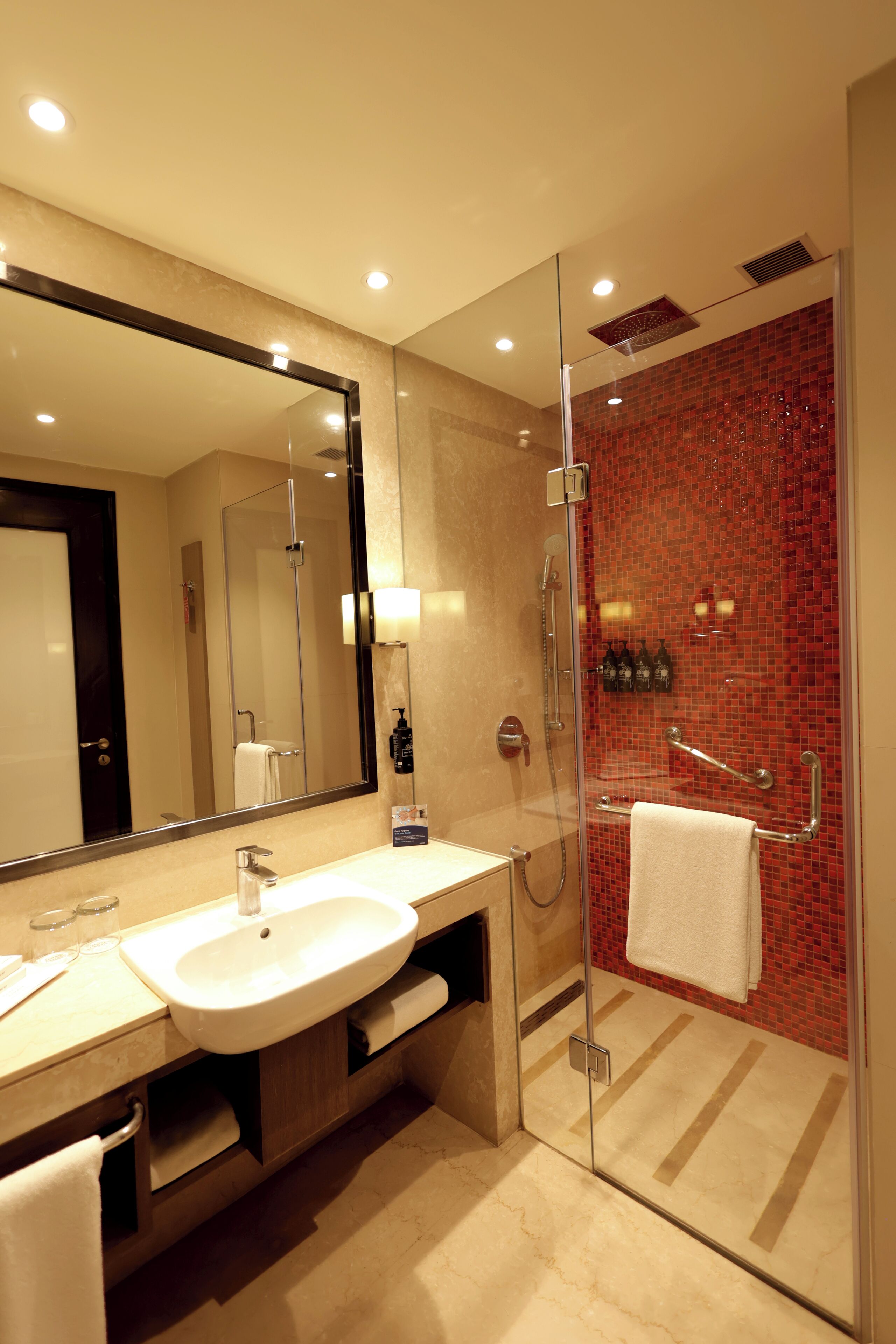 A modern bathroom with a glass-enclosed shower, a large mirror, and a beige vanity with a sink and towel rack.