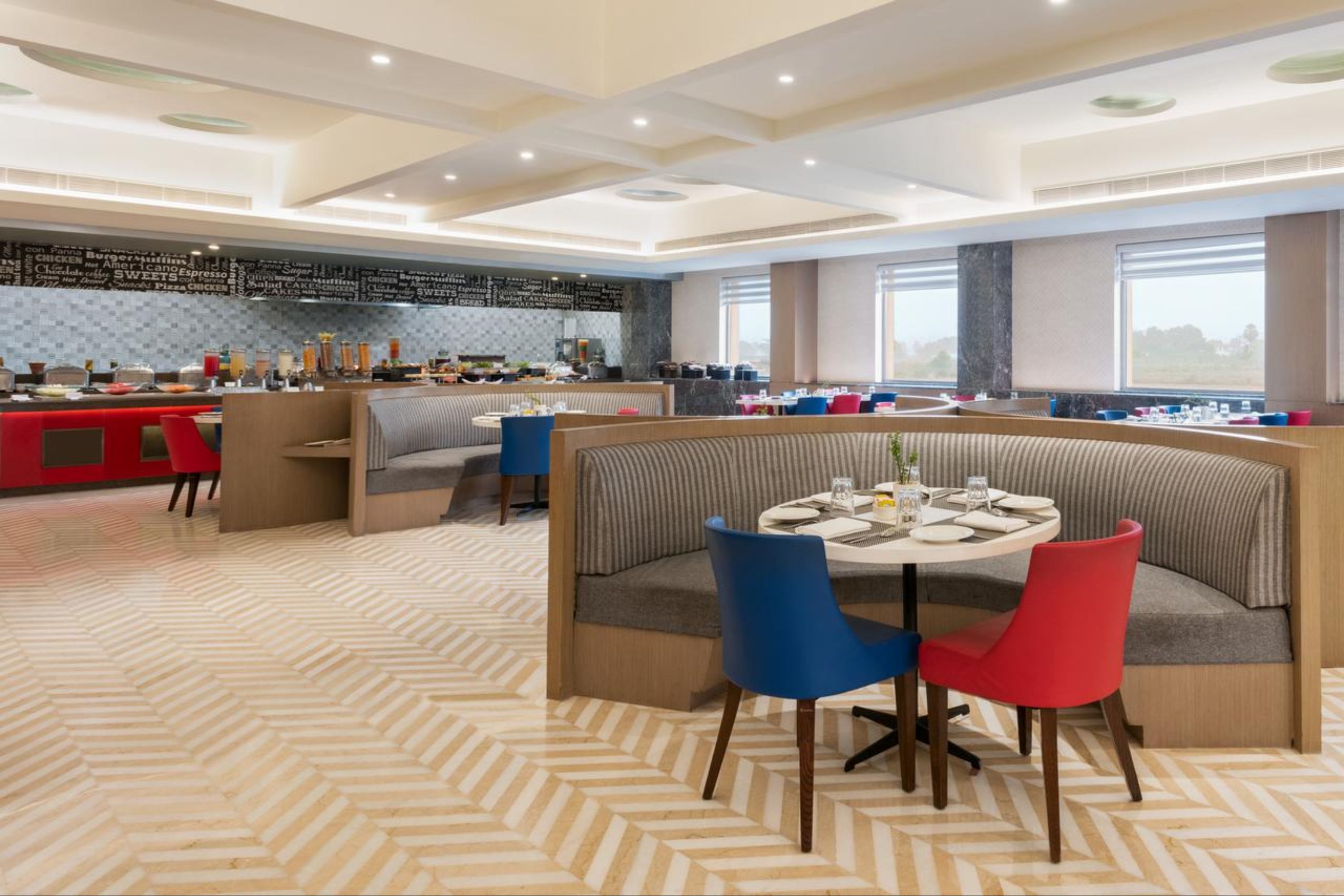 Un comedor moderno con una mesa redonda central, sillas rojas y azules, y una cocina al fondo.