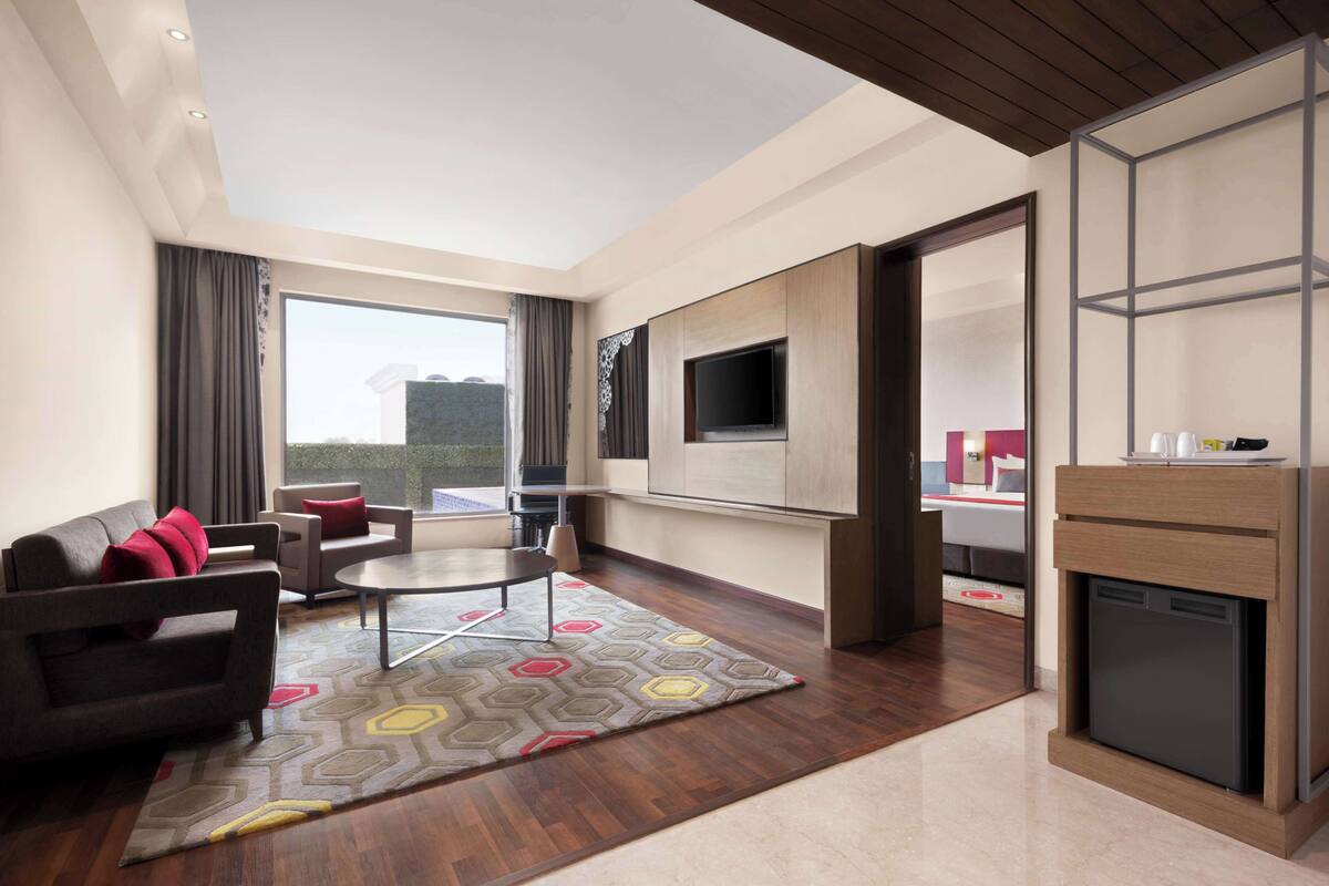 A modern hotel room with a sofa, armchair, dining table, and a built-in TV.
